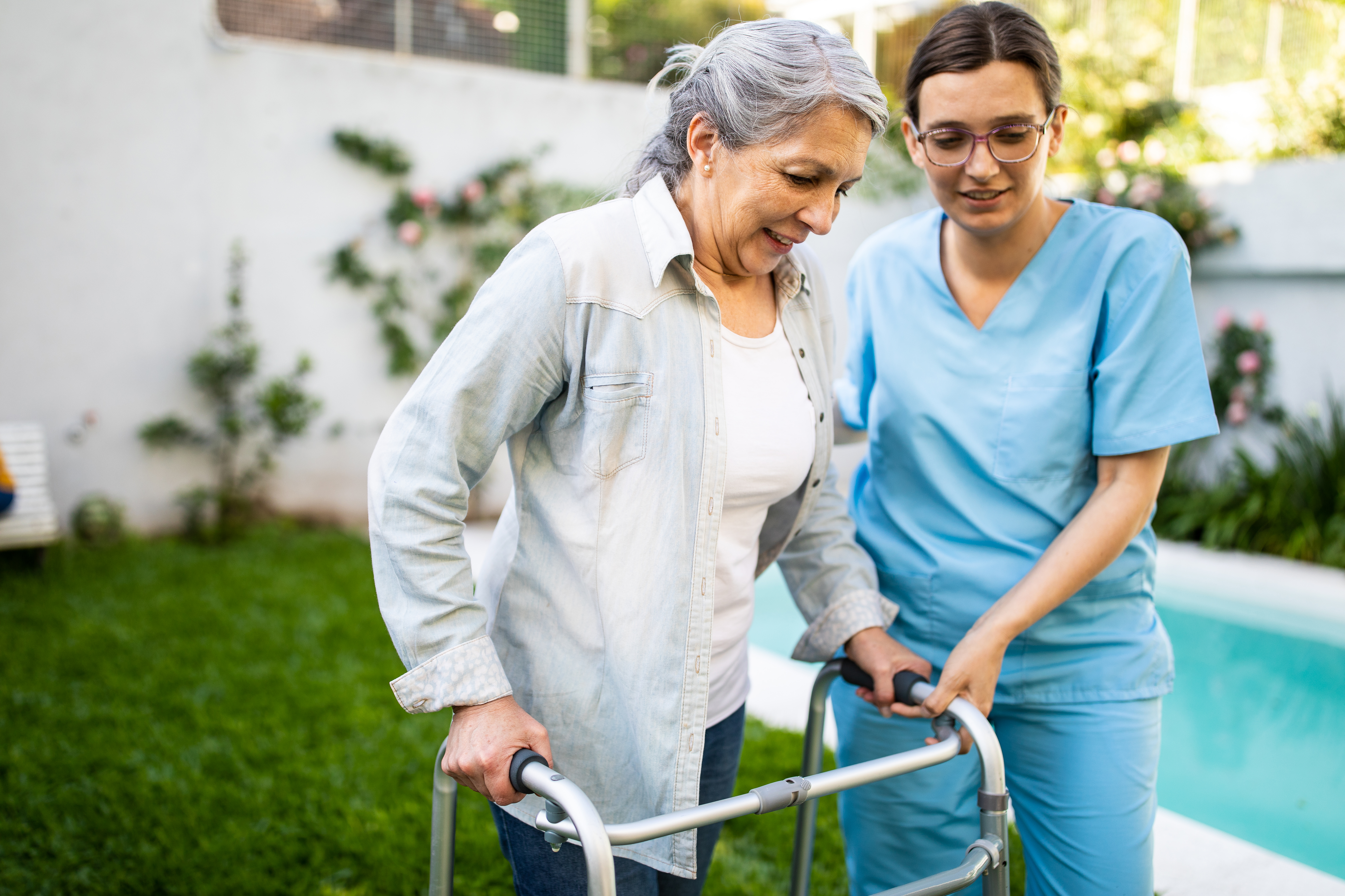 Young female nurse supporting old woman to walk outdoors with mobility walker, in retirement home